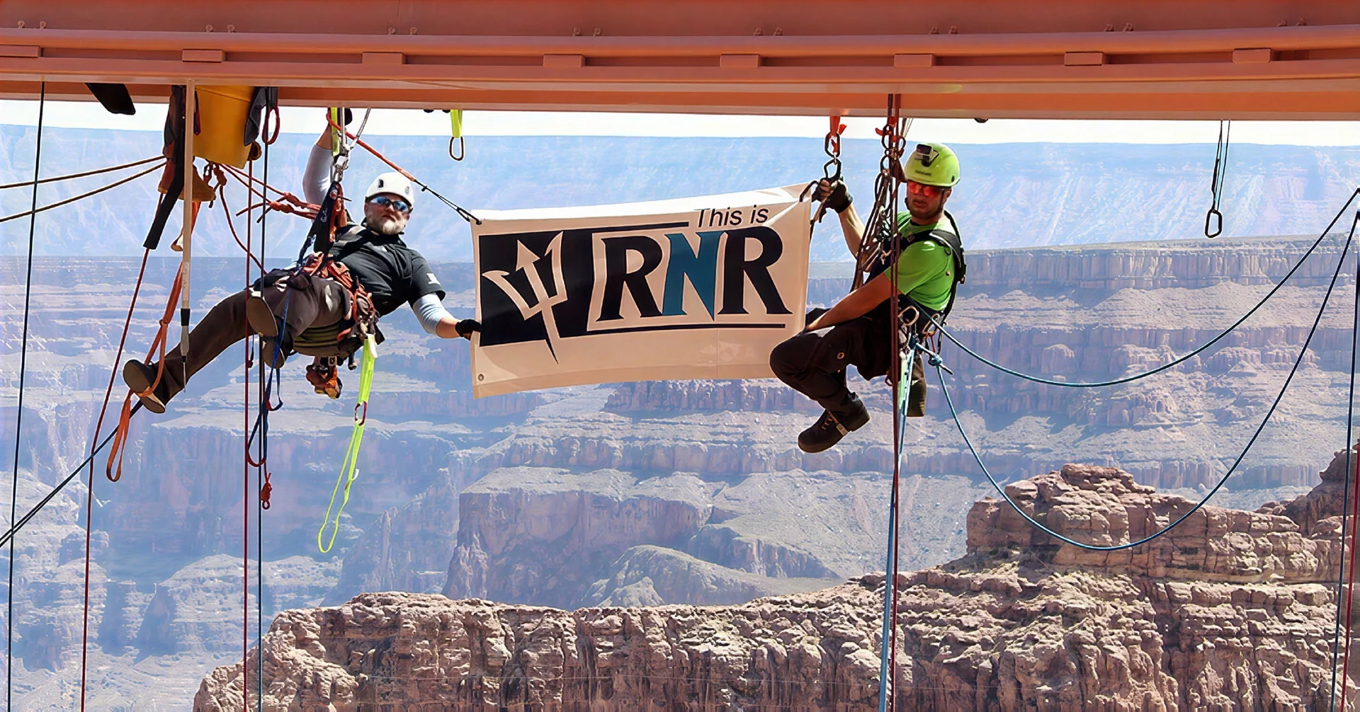 Two rope-access professionals holding an RNR banner above a canyon